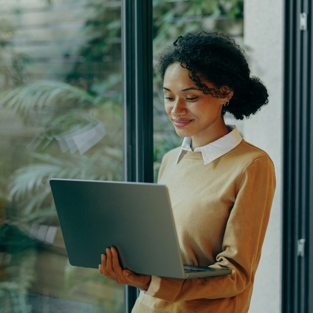 Female freelancer working laptop remotely while standing near window at home. Distance work concept